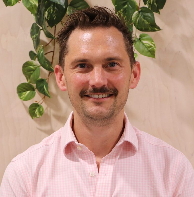 Smiling man in a pink shirt with greenery background.