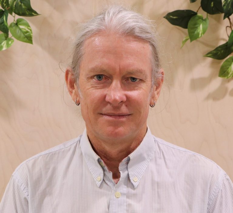 A man with white hair and white formal shirt poses in front of green plants indoors.