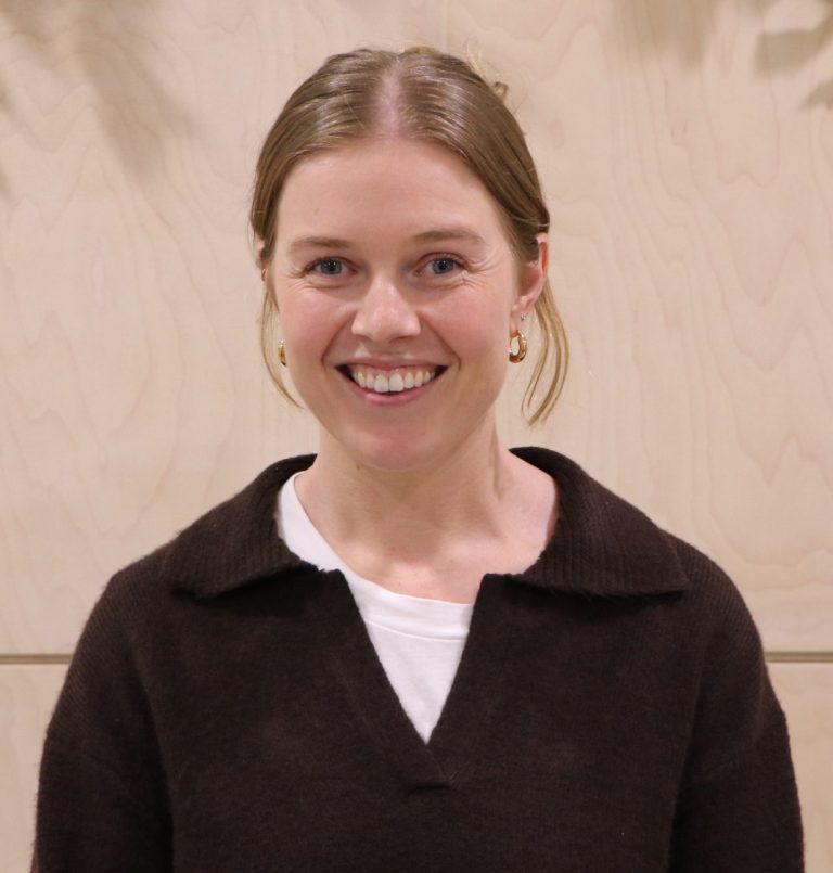 Smiling blond woman in brown sweater against a light wood backdrop.