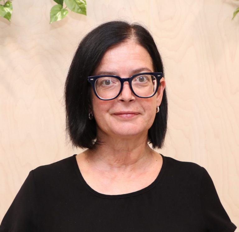 Woman with dark hair and glasses poses against wooden backdrop and plants.