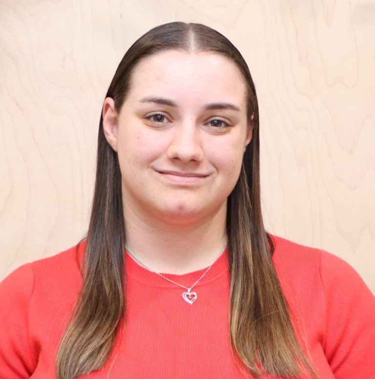 Portrait of a smiling young woman in a red shirt against a light wood background.
