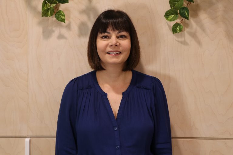A dark haired woman smiling in a blue top, surrounded by green plants.