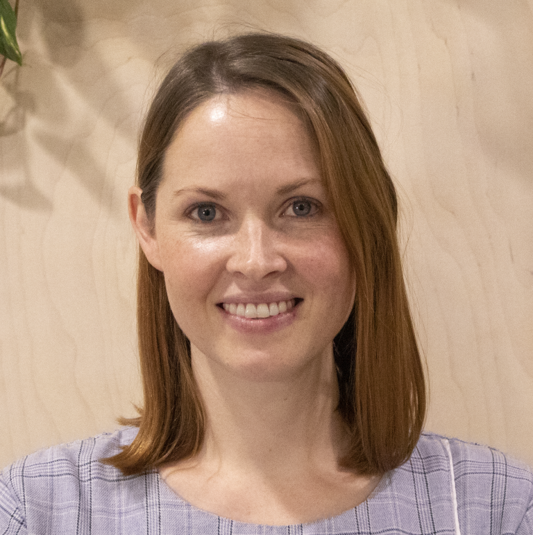 A smiling individual in a casual setting with plants in the background.