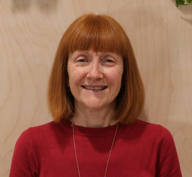 A smiling individual wearing red sweater in a casual setting with plants in the background.
