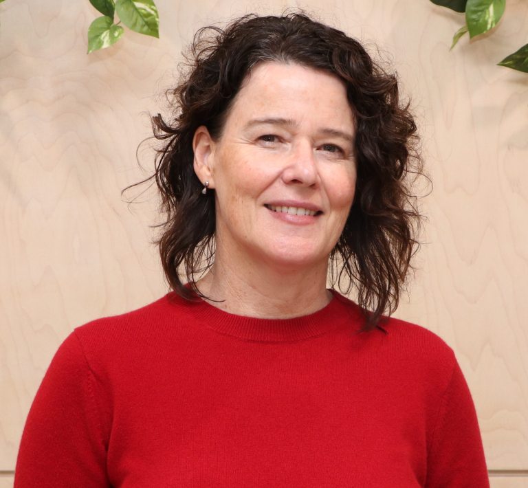 A curly hair woman smiling in front of a wooden backdrop with green plants.